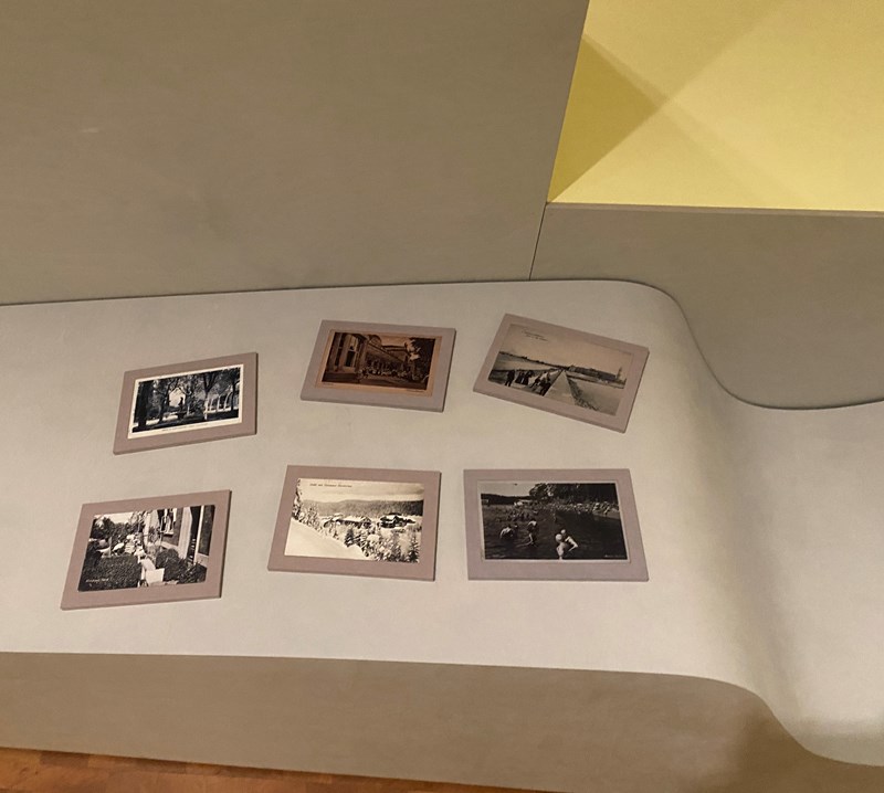 Et fotografi av seks objekter plassert på buet møbler i utstillingsrommet. Objektene er reproduksjoner av gamle postkort på små håndholdte tavler. Postkortene viser ulike kurbad, badebyer og andre destinasjoner i Europa som er knyttet til helse og velvære.