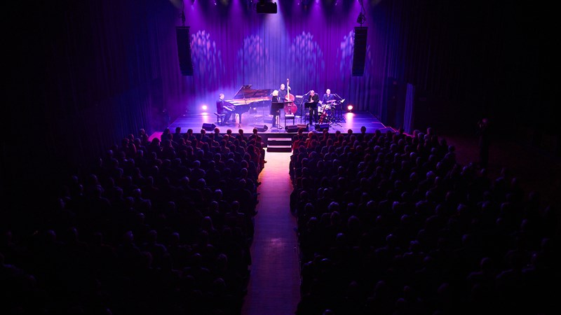 En fullsatt festsal omgitt av mørkt rosa lys, og spotlys mot scenen, hvor en gruppe jazzmusikere skimtes.